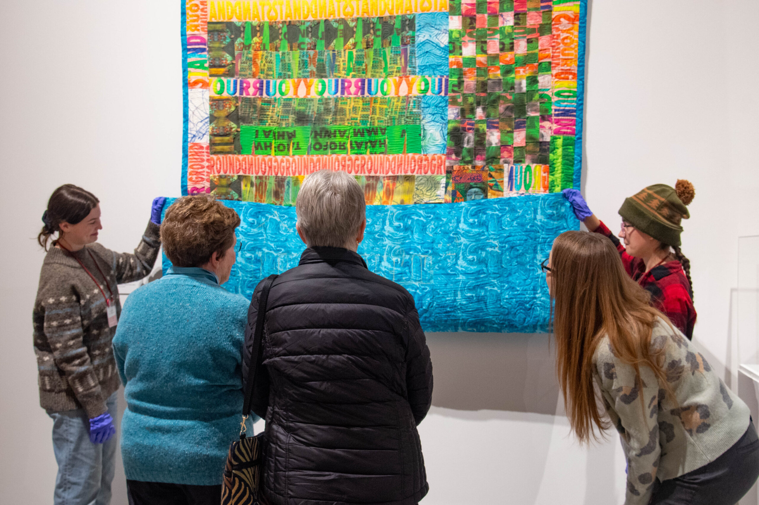 Museum visitors observe a quilt held up by museum student staff.
