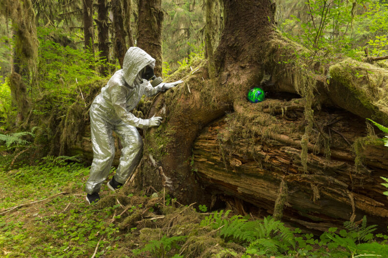 woman in hazmat suit looking at a glass orb in a forest of trees and moss