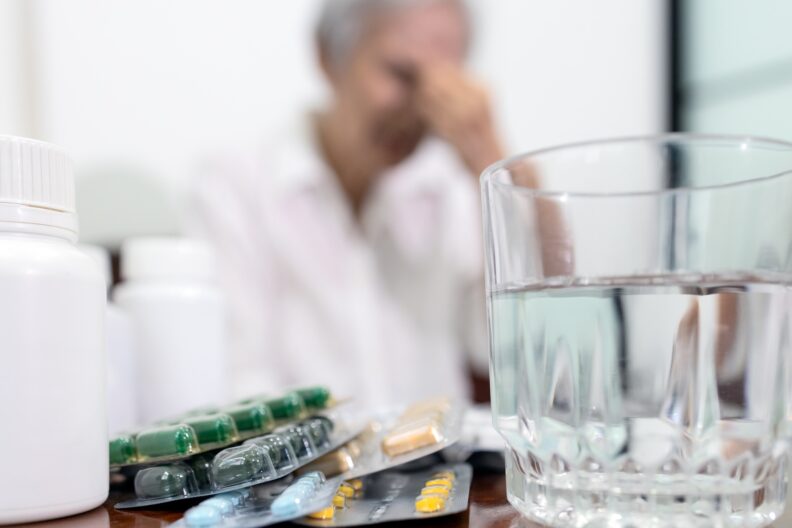 Photo displaying drugs,medicine bottle,glass of water,treatment medications to reduce pain for senior woman,old elderly in background suffering from migraine,headache with focus on tablets pills or capsules