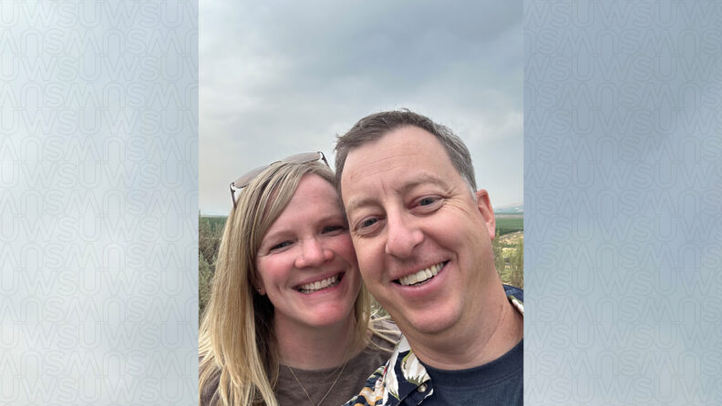 Meredith and Adam Richards smiling together in a close-up outdoor photo, with a scenic landscape of hills in the background.