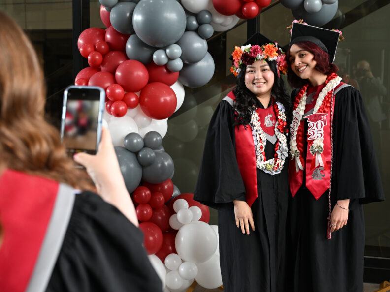 Two students in graduation regalia pose for a photo in front of a crimson and grey balloon arch at the Spring 2025 Graduation in Spokane, WA. Photo by Bob Hubner.