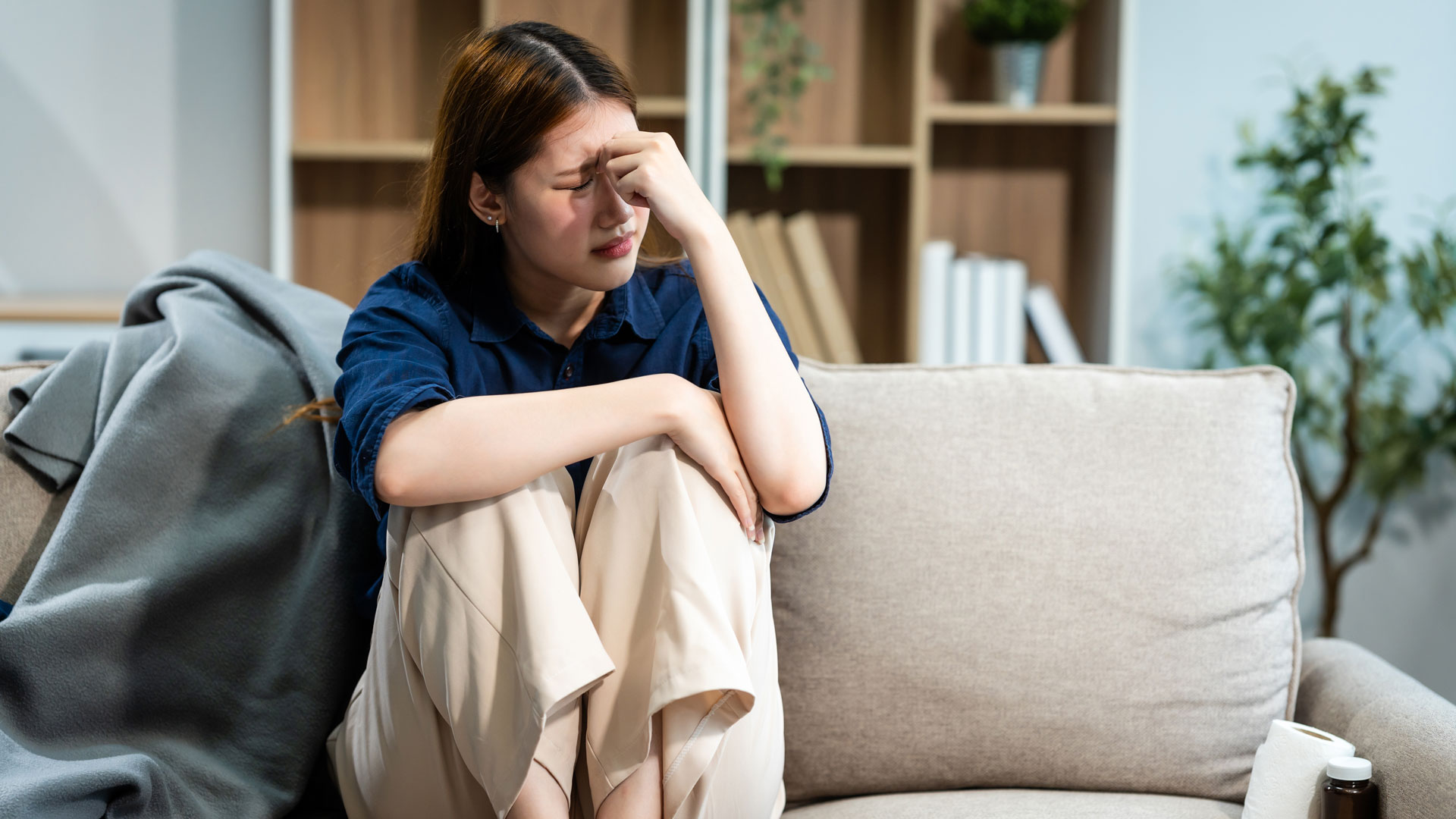 Woman sitting on a couch with her knees up in front her, holding her forehead in pain, looking upset.