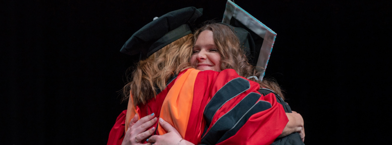 Cadence McLin and Associate Professor Theresa Bowden embrace at commencement, wearing academic regalia.