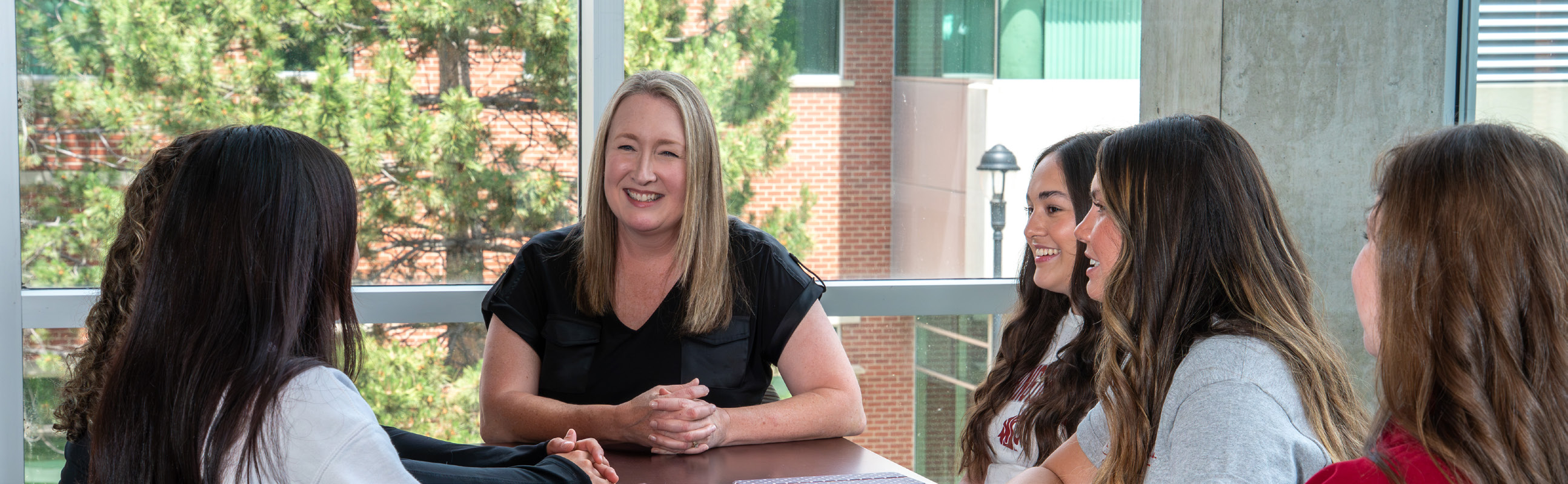 A Dean Anne Mason talks with four BSN students around a table in a bright campus gathering space.