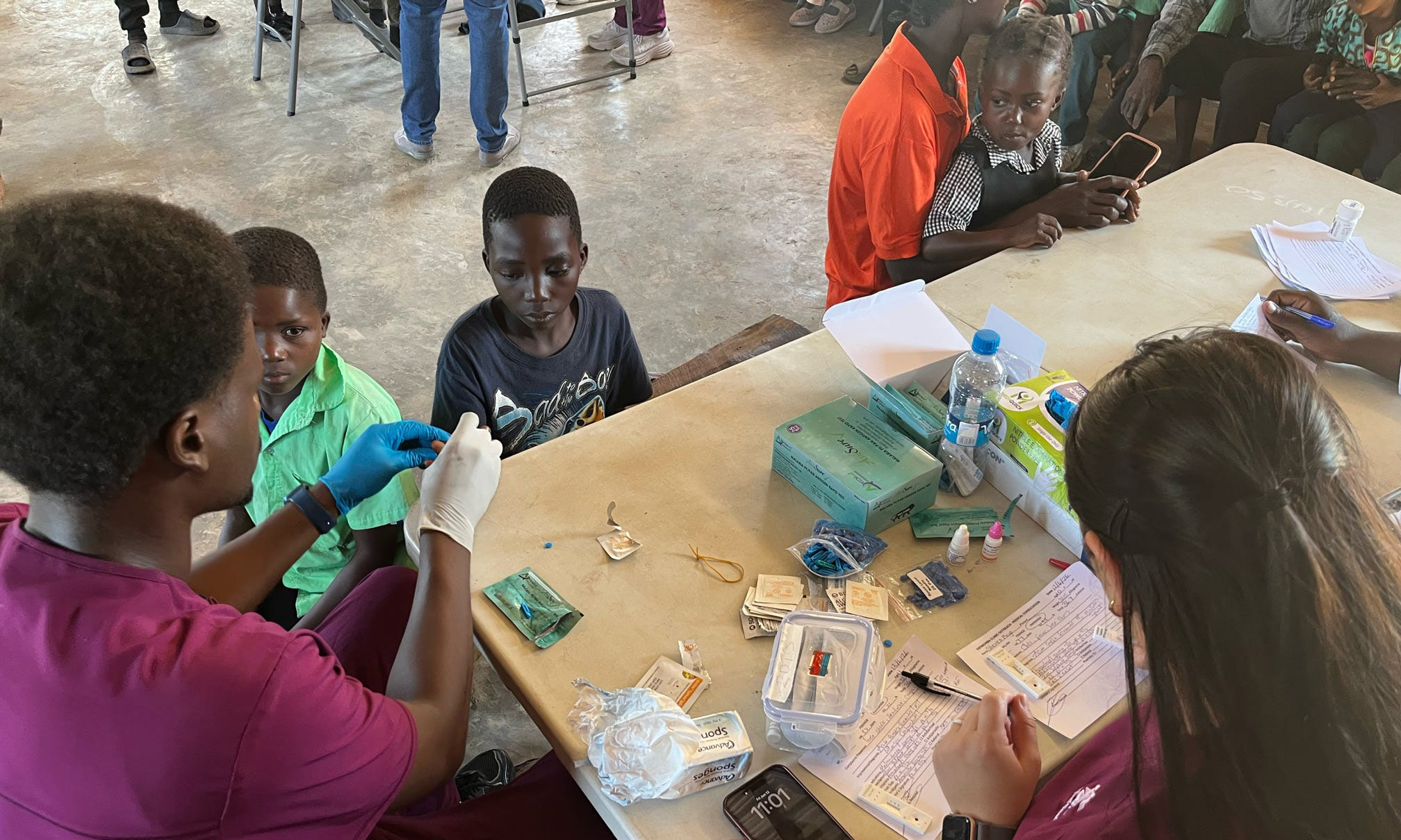 WSU nursing students Nasir Hussein and Abby Elliot prepare blood samples at a table with children seated nearby at a pop-up clinic in Kayenda, Zambia.