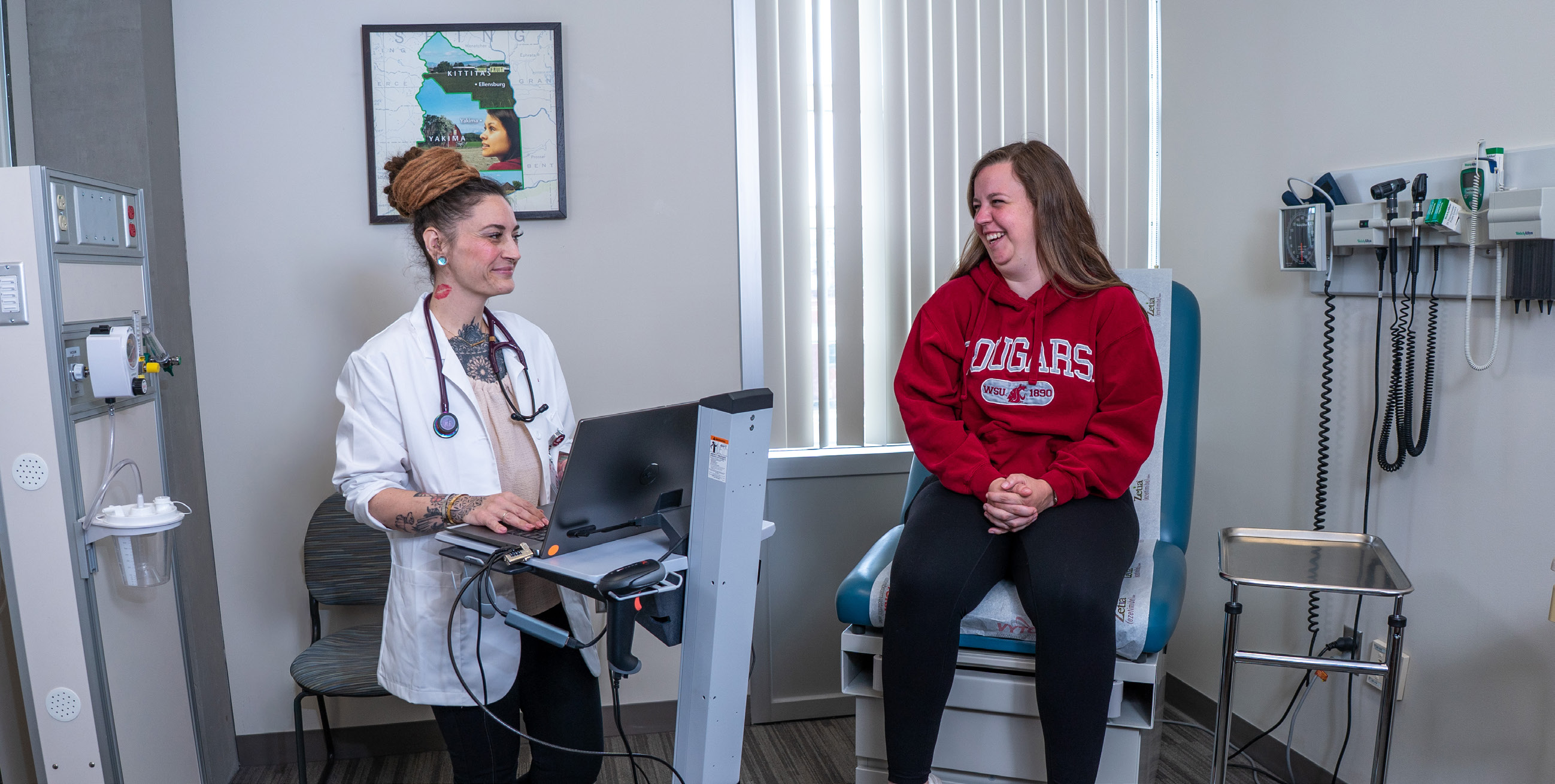Shaena Nisito and Bryn Killion practice patient care in a mock exam room; one sits at a laptop wearing a white coat and stethoscope while the other sits on the examination table.