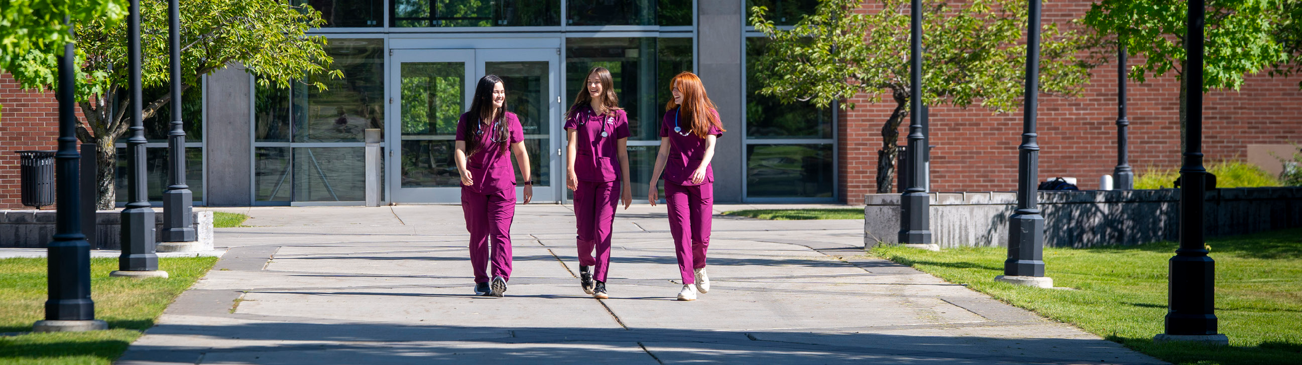 Three people in crimson scrubs walk together along a campus path outside a brick building.