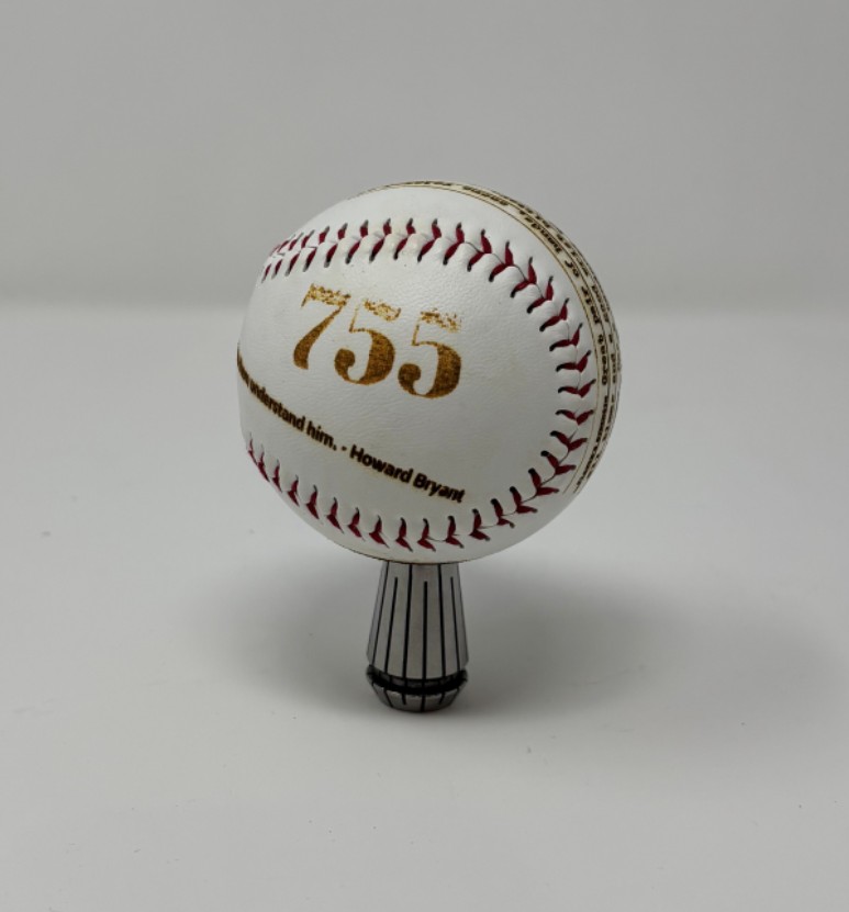 A baseball with red stitching and the number 755 is sitting upright on a stand. There are additional laser printed markings from a class project that are partially displayed.