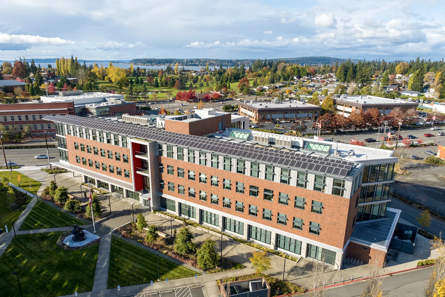 Aerial view of the WSU Everett campus looking northwest on a sunny autumn day. The changing colors of the trees within the Evergreen Arboretum & Gardens stands out in the background of the scene.