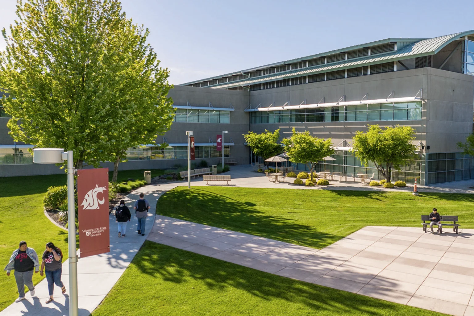The WSU Tri-Cities campus in front of the Consolidated Information Center looking northwest on a sunny day. Students are walking to and from the building and one student is seated at a park bench.