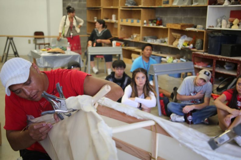 Dr. Brigman sews a canvas skin to a canoe frame while youth watch in the background.
