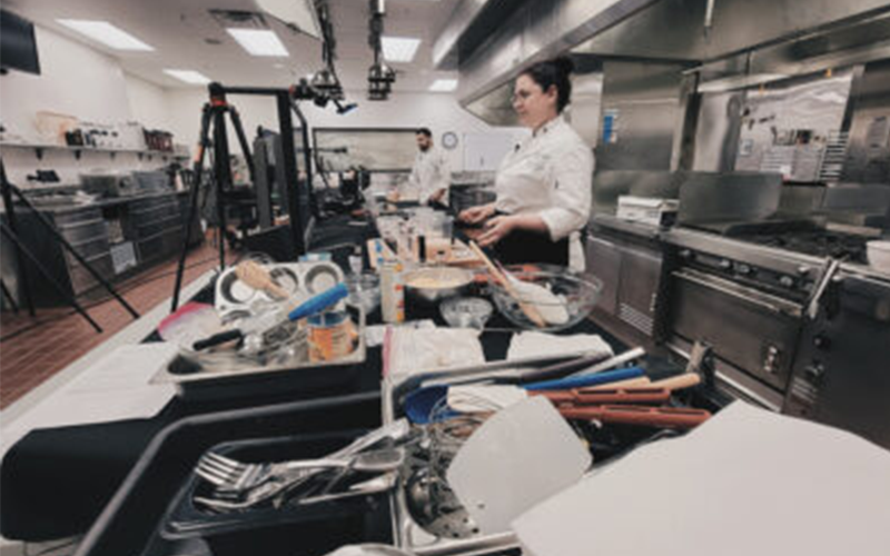 A chef in the School of Hospitality Business Management speaks to a camera as they conduct a virtual cooking class in a professional kitchen.