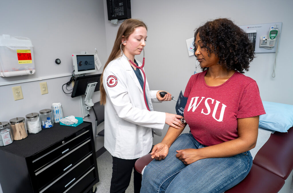 First-year medical student with a standardized patient in a WSU shirt at the Virtual Clinical Center. The student is measuring blood pressure.