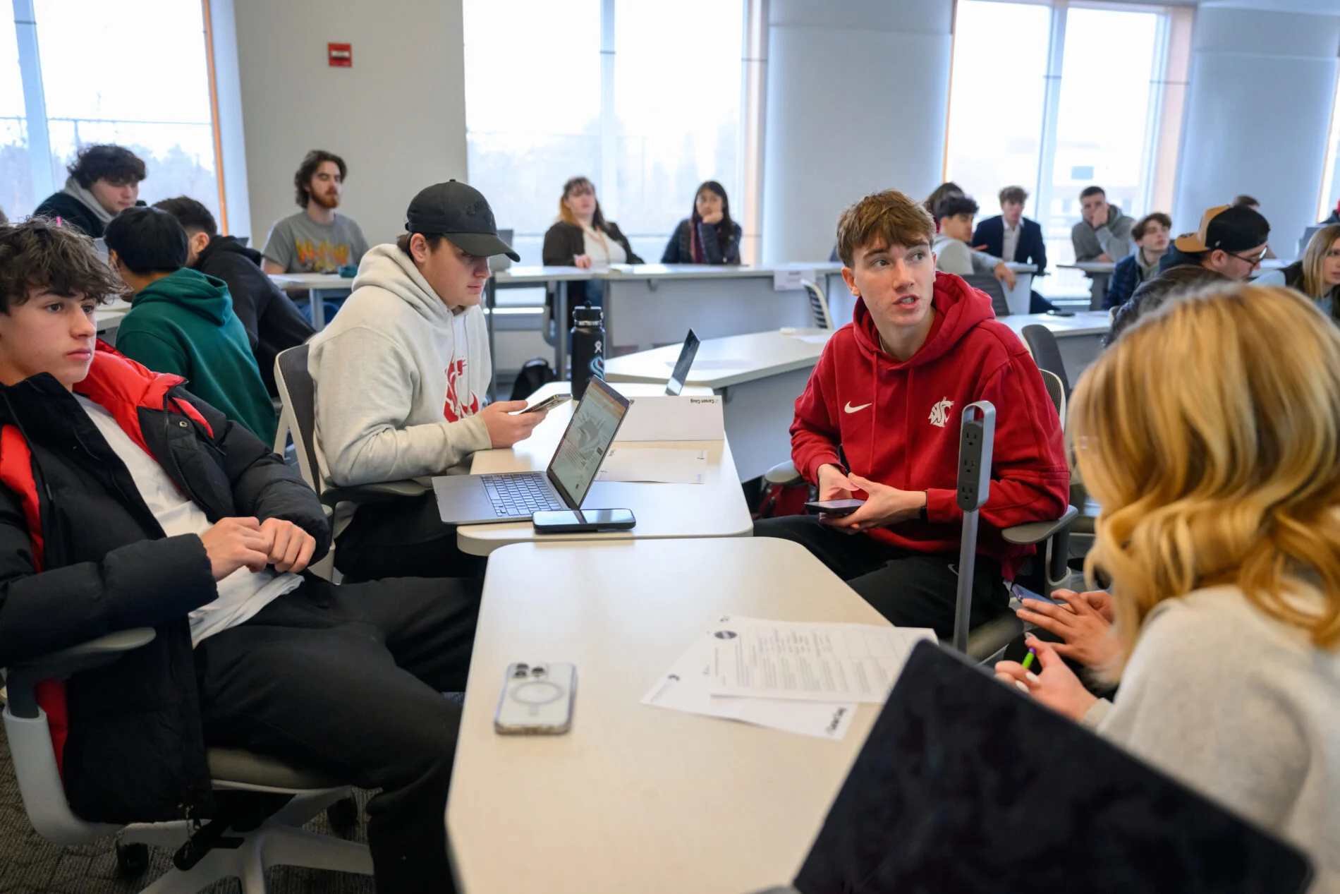Several undergraduate students engage in a group discussion during a lecture. They are using laptops, mobile phones, and printouts.