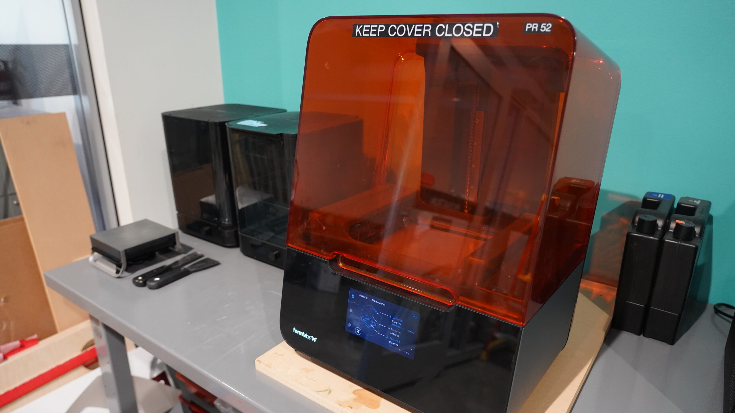 A 3D resin-based printer on a table. Two resin tanks are shown along side. A small wash station and curing station on shown on the far side of the printer.