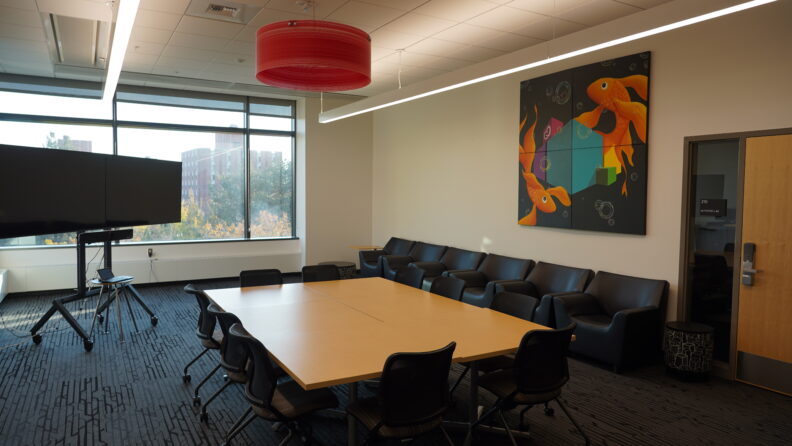 A room with comfortable chairs along the wall, large table in center with chairs. Two flat screen displays stand at end of the table. A colorful painting is hung on the wall with a red round chandelier hanging from the ceiling.