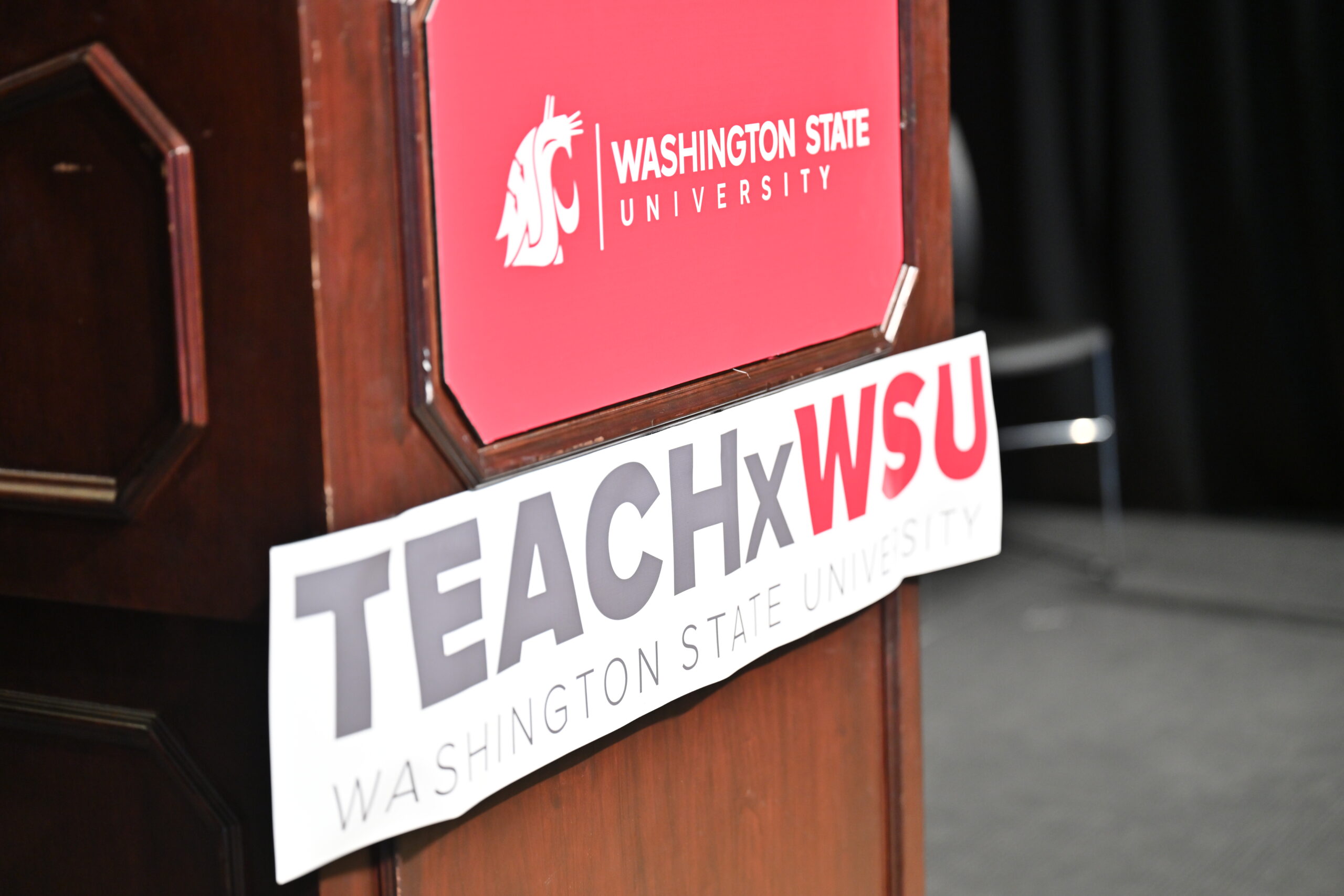 A wooden podium featuring a crimson sign with a white Washington State University logo, and underneath a white sign reading TEACHxWSU.