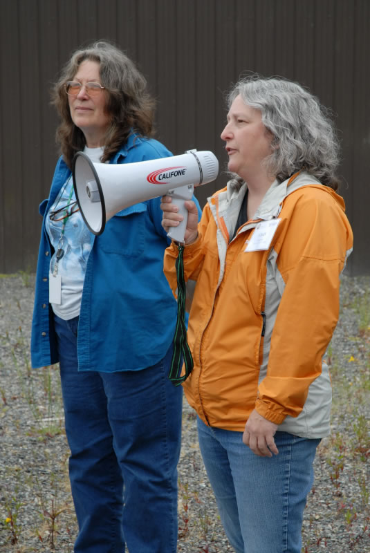 two women standing in gravel, the one on the right in an orange and light grey windbreaker coat has a megaphone. the one on the left is wearing dark blue jeans and a slightly lighter blue denim button-up shirt unbuttoned. She has a badge and glasses hanging around her neck.