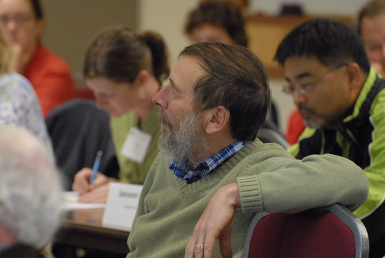 a man faces the left with his arm on the back of the empty chair next to him