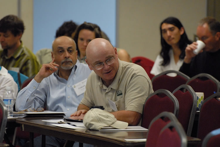 Two men at a table, one smiling and talking and the other sitting back and listening