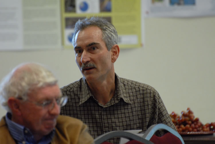 A man in a plaid shirt having a conversation with someone unseen. A man in a brown jacket blurry in the foreground