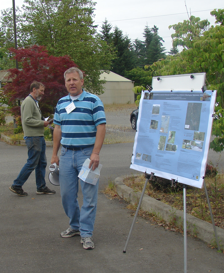 a man holding a map next to a scientific poster on a tripod stand. another man walks behind him, consulting some papers.