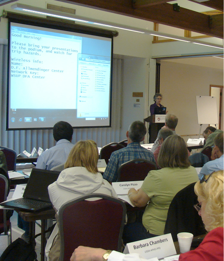 people at tables with nametags in front of them, facing a lady speaking at a podium and a screen with instructions for presenters