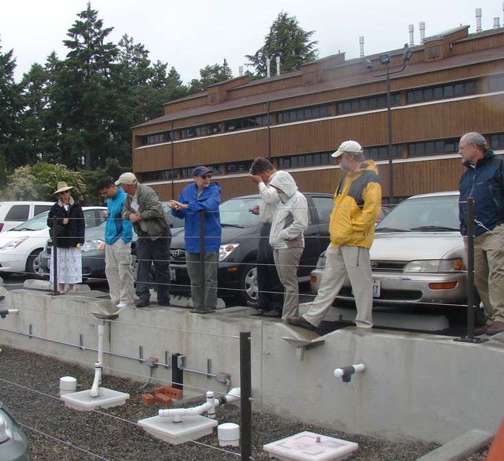 four men standing behind a fence made of four horizontal wires in the parking lot of a brown building, raised above the ground with a cement wall that has pipes coming out of it and divots to encourage water collection