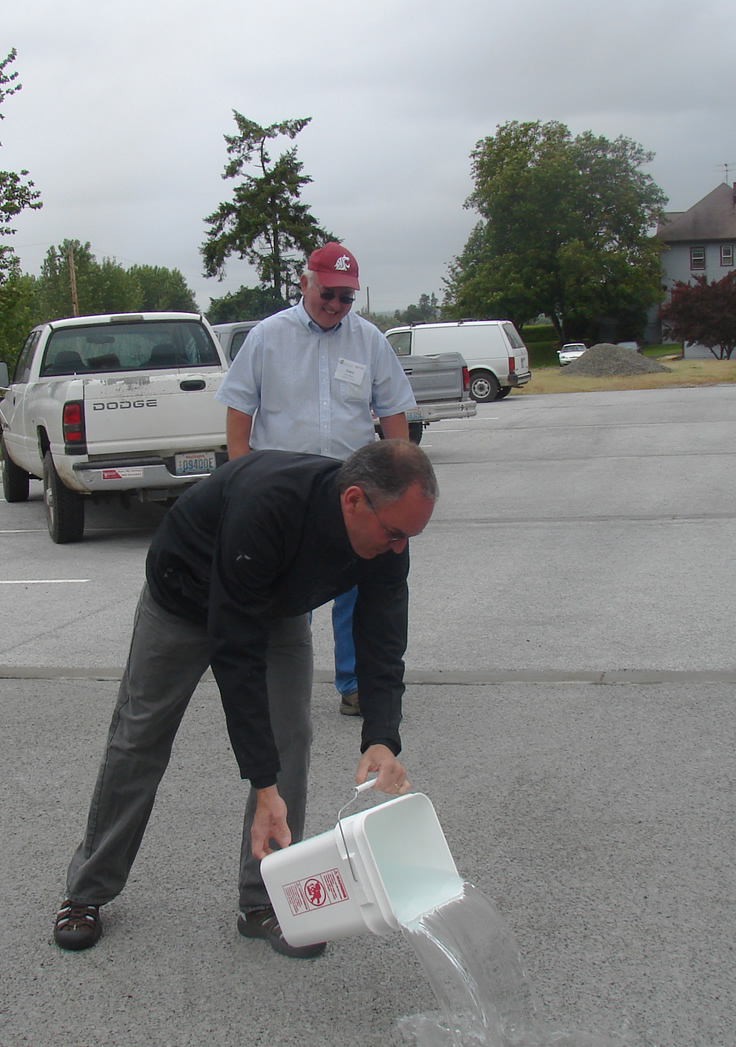 a man pouring a bucket of water onto a parking lot with a man standing behind him smiling