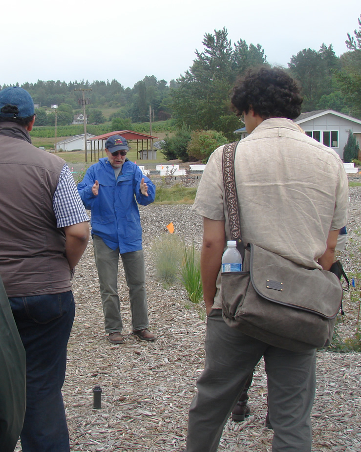 two men in the foreground, one man facing them and speaking, all standing on wood chips with some bunched grasses
