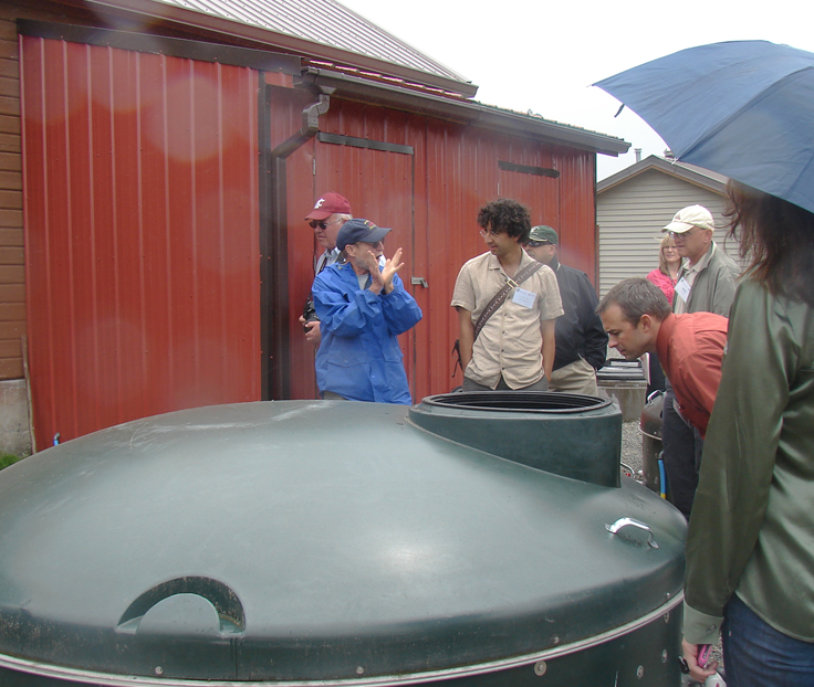 A man talking, using his hands to demonstrate. A man listening. Two men behind them walking past. A man in front of them bent over an opening in a large green container.