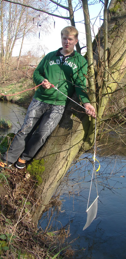 A male in a green sweatshirt and black jeans with decorations of wings on the front of the legs down to his knees. He is leaning against a tree on the creekbank, holding the rope attached to a bait bag, which dangles down out of the frame.