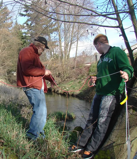 Two males on a streambank. One holds a thin red rope that leads to the hand of the other man. It attaches to a thinner white rope strung along to his other hand and on the end of the rope can be seen a bait bag labeled with a yellow flag.