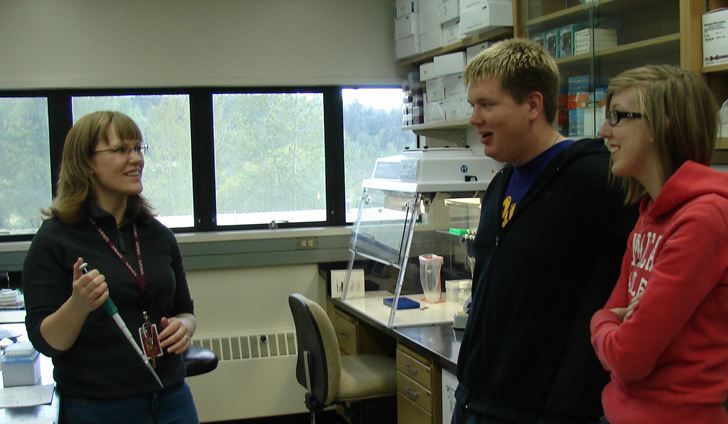 A woman wearing a WSU badge holds a pipette in her right hand. Facing her, two students smile.