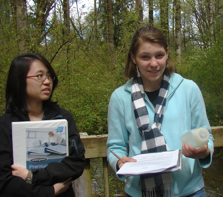 Two students standing in front of a wooden railing. The student on the right is holding stapled papers and a spiralbound notebook in one hand and a small square bottle in the other. The student on the left is holding a binder.