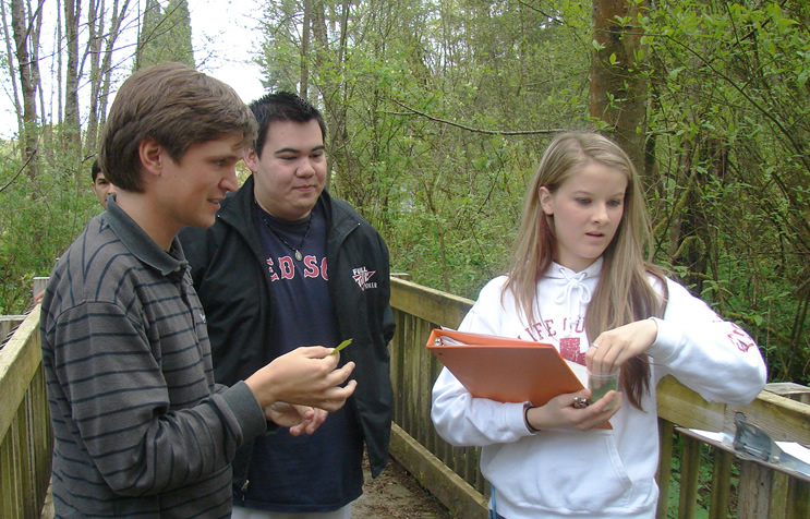 Three students stand facing toward the right of the frame.. The student on the left is holding a leaf out to the third student while the second student watches. The third student, on the right, holds an orange binder and is reaching her hand into a small clear container with something green in it.