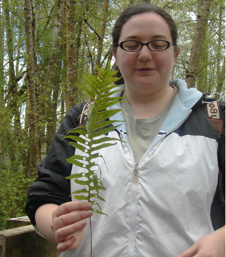 A student with glasses holds up a sword fern with heart-shaped holes punched in some of the leaves.