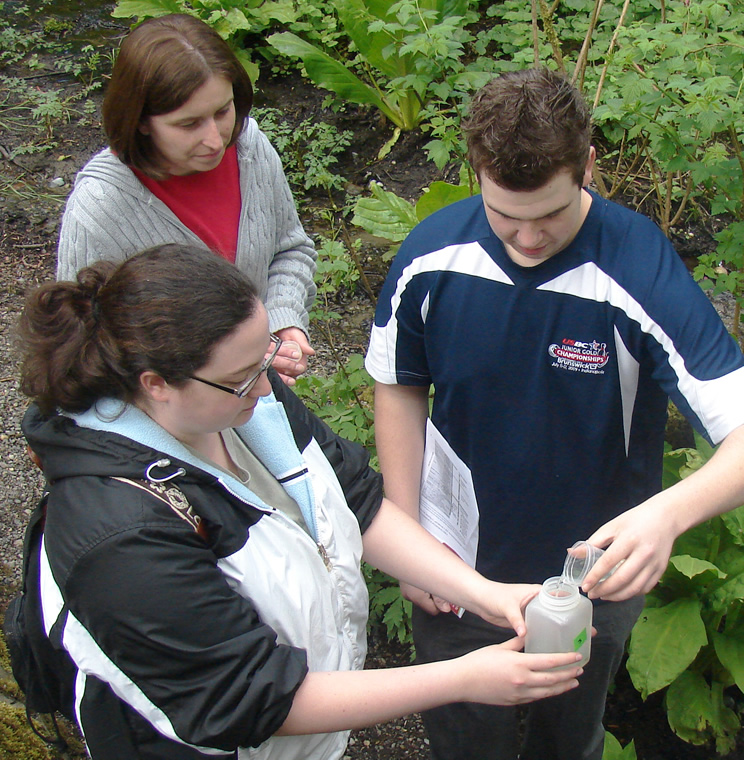 Two students stand together while a third watches behind them. The student on the left holds a small square bottle with no lid in both hands while the student on the right pours the contents of a small clear container into the bottle.