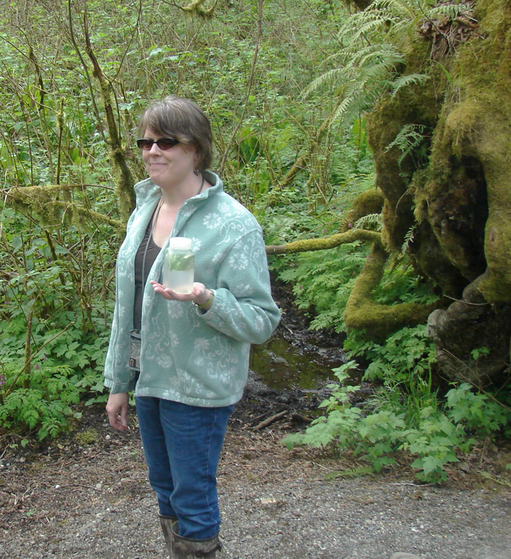A student in sunglasses holds one palm face up with a small square bottle resting on it. The bottle has water in it, and green things is floating towards the top.