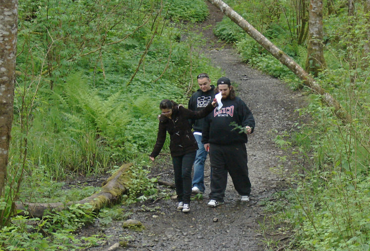 Three students walking on a rocky dirt path with lush green foliage on either side. The student in front is holding up some papers toward the second student with a small red and black device held in the same hand behind the papers. The third student is just walking behind them.