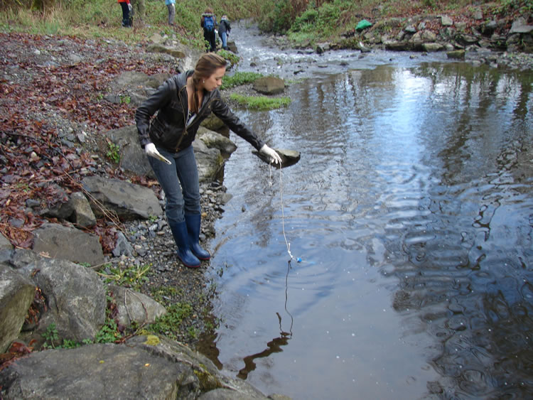 A girl standing on the banks of a creek, holding a thin white rope attached to a small clear tube with a blue lid just below the water. The water in this area of the creek is fairly smooth with just a few ripples, but at the top of the frame it can be seen that there is some slightly rougher-looking water flowing over some rocks.