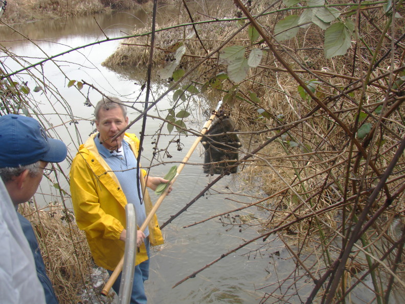 A man in a yellow jacket stands in the water, holding a yellow pole with several bait bags hanging from it.