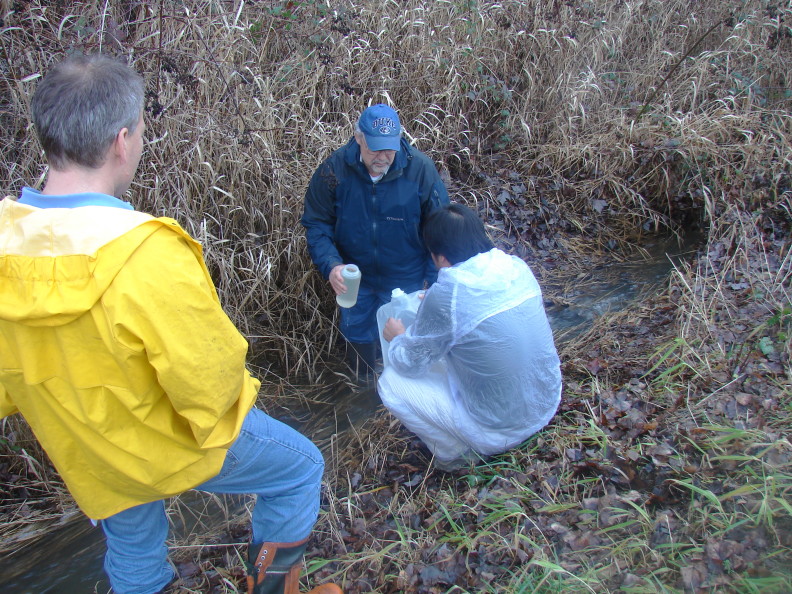a man in a blue hat standing in a ditch with flowing water and holding a Nalgene with the top off. Squatting in front of him a student in a white jacket holds a water bag. A man in a yellow jacket faces them.