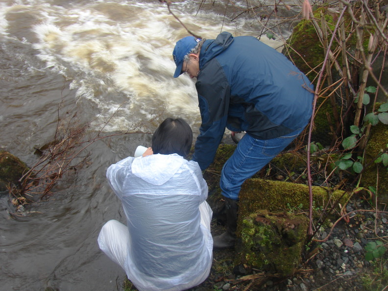 A man in a white coat squats by the side of a fast-moving creek, apparently pouring something with his left hand. A man in a blue coat stoops over next to him, holding something out of sight behind the other man's torso.
