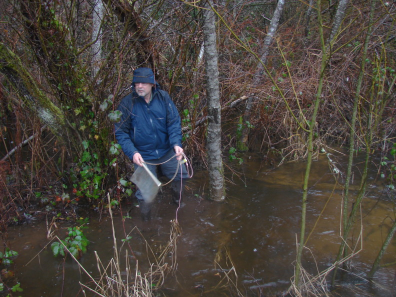 A man in a blue jacket stands in brown water with trees growing in it. He holds in his hands a bait bag with its rope attached and dangling down.
