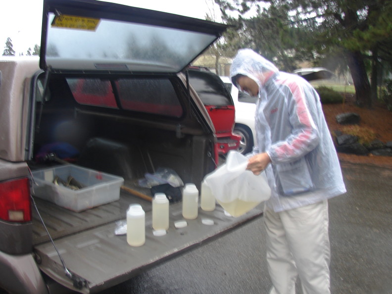 A male standing by the open back of a brown truck. He is pouring water from a bag into open nalgene containers with their lids next to them.
