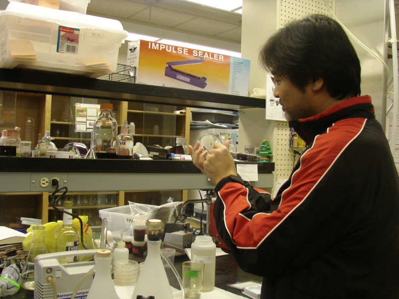 A man in a black and red jacket stands by a black lab bench next to the filtration bottle, using a pair of tweezers to place the round filter from the bottle onto a petri dish.