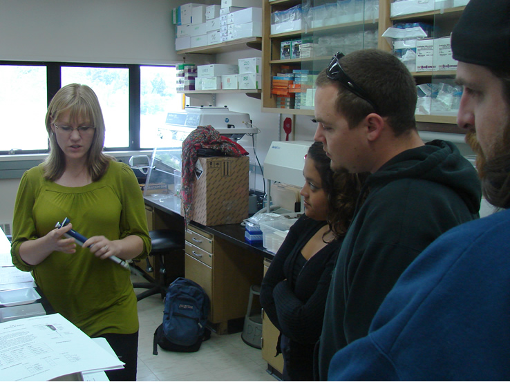 a woman in a green shirt holds a pipette with a disposable tip attached and looks at it as two students watch