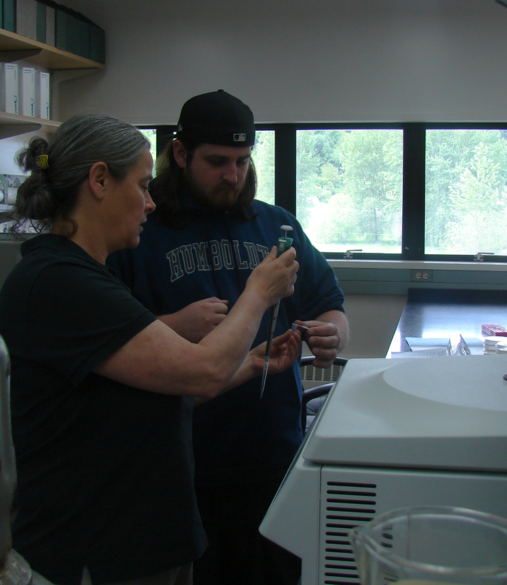Two students standing in front of a midsized centrifuge with its lid down. The student closer to the camera is holding up a pipette with a tip on it as the second student hands them a 2mL microcentrifuge tube with an open pop-up lid.