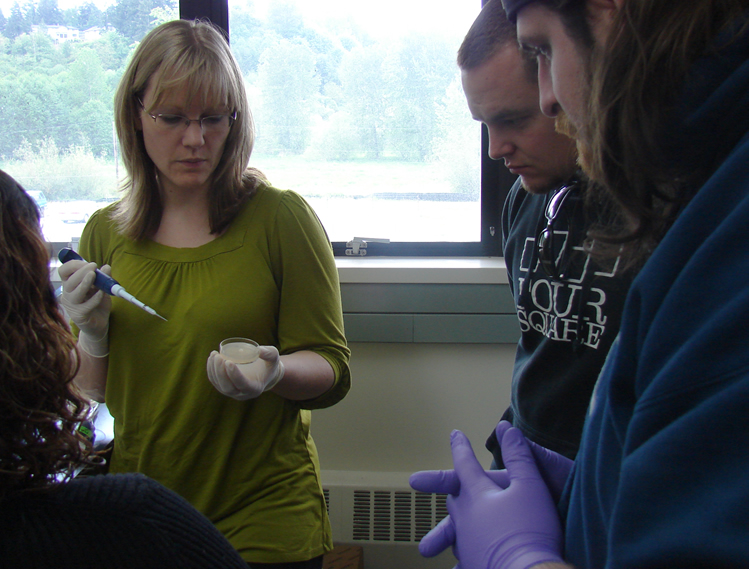 a woman in a green shirt wearing latex gloves is holding a small petri dish without its lid on and a pipette with a tip pointed at it as two students watch. The student closer to the camera is wearing purple gloves.